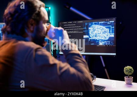 Programmer drinking coffee and doing maintenance on artificial intelligence neural networks computerized model. Tech support expert enjoying hot beverage while checking up on AI systems Stock Photo