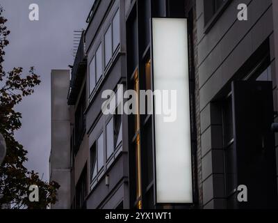 A vertical rectangular white sign glows at dusk on a German commercial street, offering a blank, illuminated space against building facades. The brigh Stock Photo