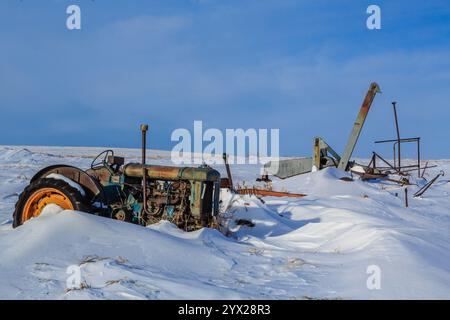 An old tractor is sitting in the snow. The tractor is rusted and has a broken front wheel. The tractor is surrounded by snow, and the sky is clear. Th Stock Photo