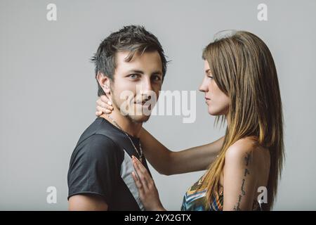 Young couple embracing each other while smiling and gazing at the camera, posing for a cheerful portrait against a soft gray background, radiating int Stock Photo