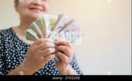 Happy older Asian woman holding a lot of Thai money that she has won, feeling excited. Retirement concept. Stock Photo