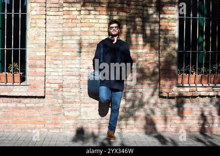 A Caucasian man in a navy coat, jeans, and boots leans casually on a brick wall. He wears sunglasses and headphones, carrying a bag. Urban, stylish vi Stock Photo