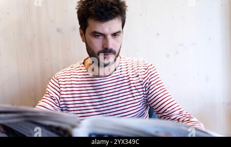 Caucasian man with a beard wearing a striped shirt reads a newspaper indoors. Focused expression in a minimal wooden interior setting. Stock Photo