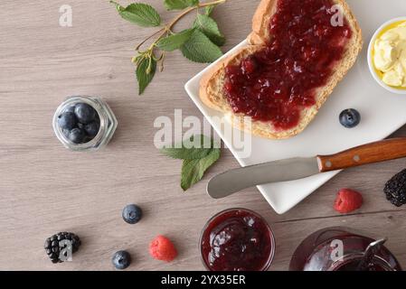Toast with forest fruit jam on a white plate on a wooden bench with jars and berries. Top view. Stock Photo