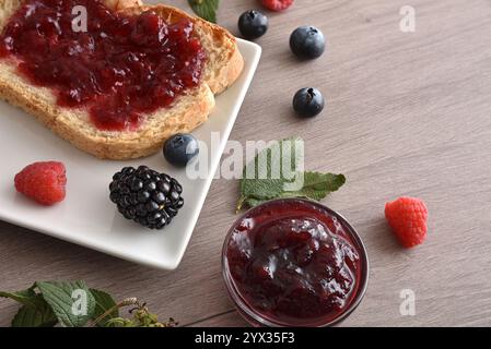 Detail of toast with forest fruit jam on a white plate on a wooden bench with jars and berries. Top elevated view. Stock Photo