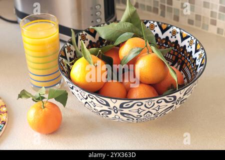 Clementines, mandarins with leaves in ceramic bowl. Fresh juice of citrus fruits in glass on table in kitchen. Mosaics and pattern on dishes.  Stock Photo