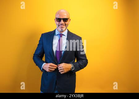A man in a dark blue suit stands in front of a yellow wall. He buttons up his jacket. Stock Photo