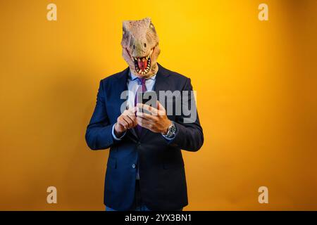 A man wearing a dinosaur mask using a smart phone on yellow background. Stock Photo