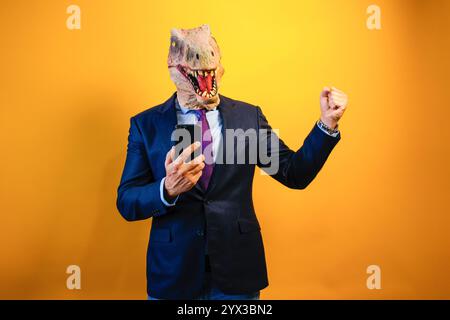A man wearing a dinosaur mask using a smart phone on yellow background. Stock Photo