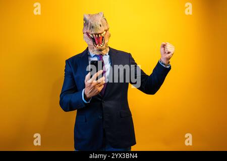 A man wearing a dinosaur mask using a smart phone on yellow background. Stock Photo