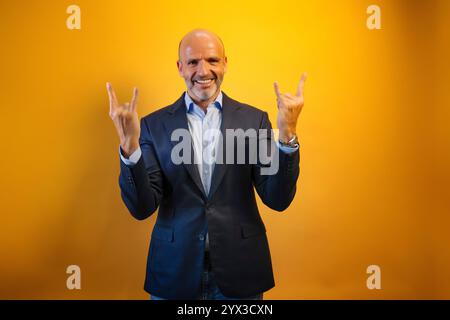A man in a suit raises his horns with his hands in front of a bright yellow wall. Stock Photo
