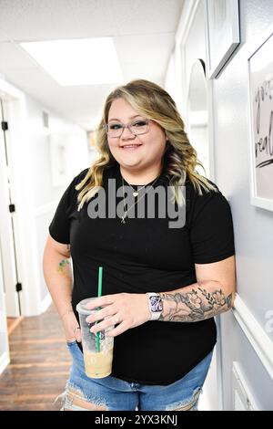 Woman in black shirt, holding iced coffee, smiling in hallway Stock Photo