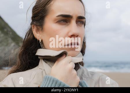 Close-up of a young woman observing the ocean while adjusting her coat on a chilly day. Stock Photo