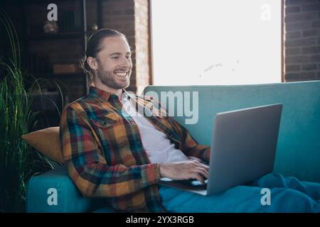 Young man relaxing with laptop on a sofa in a stylish loft living room, enjoying leisure time and smiling during a sunny day at home. Stock Photo