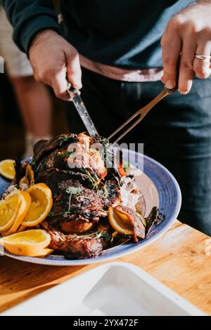 A person is cutting a large piece of meat on a blue plate. The plate is topped with oranges and other vegetables Stock Photo