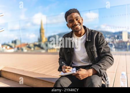 Happy african man writing notes sitting on a terrace with city views in a sunny day Stock Photo
