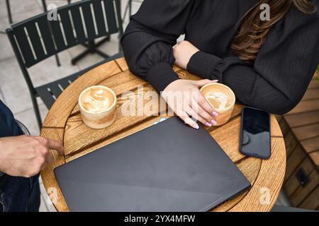 Two people having coffee outdoors, enjoying lattes, with a laptop and smartphone on the table, casual interaction concept Stock Photo