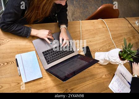 Top view of individuals using laptops and writing in notebooks on a wooden table, collaborative office workspace, teamwork environment concept Stock Photo