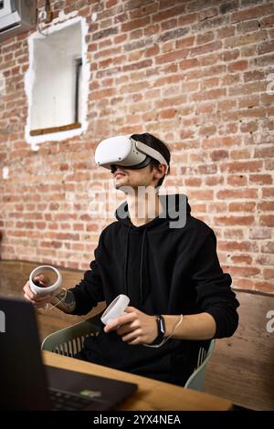 Individual in black hoodie exploring virtual space with headset and controllers sitting in a contemporary room virtual experience concept Stock Photo
