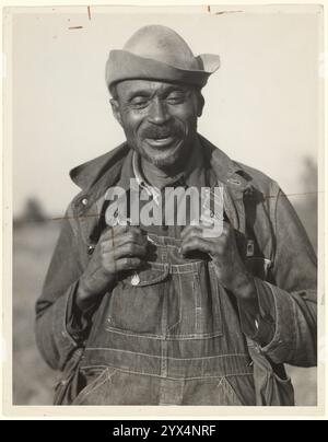 Father of Twenty-three Children, Gee's Bend, Alabama, 1937. Portrait of a man wearing a hat and denim overalls smiles at the camera. He holds the straps of his overalls at the shoulders. Stock Photo