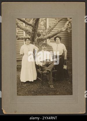 Portrait of a man and two women, about 1890-1900.  Additional Info: Portrait of a man and two women. The man is wearing a boater hat and seated in a chair at the base of a tree. He is holding a fiddle in his lap. The women are standing on either side of him, both leaning against the tree. Stock Photo