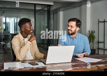 Two busy happy diverse men working on laptops discussing project at work. Stock Photo