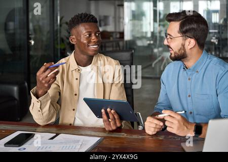 Two happy busy diverse coworkers using tablet working talking in office. Stock Photo