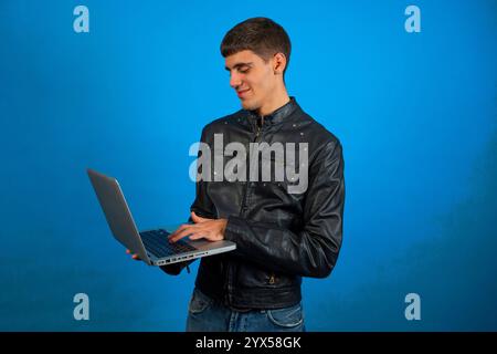 Young programmer wearing black leather jacket is using a laptop and smiling, on blue background Stock Photo