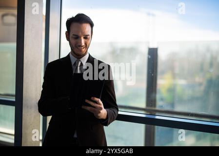 Confident young businessman in sleek suit browsing on tablet in modern office, creating professional atmosphere with cityscape view Stock Photo
