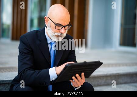 Confident mature businessman in suit browses digital tablet outside corporate office, embodying successful executive in modern city Stock Photo