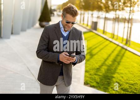 A professional man in stylish attire leisurely walks outside a sleek building, smiling as he interacts with his phone under the warm afternoon sun, ex Stock Photo