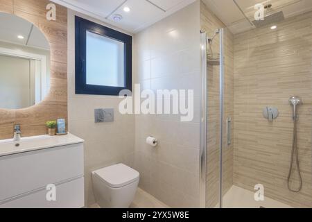 Contemporary bathroom featuring a sleek walk-in shower, minimalist vanity with a round mirror, and natural light from the window. Stock Photo