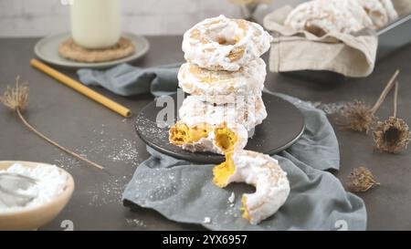 A stack of ring-shaped donuts dusted with powdered sugar, with one half-eaten donut showing a golden filling. Stock Photo