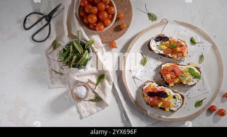 Fresh bruschetta topped with cherry tomatoes, pineapple, and herbs on a white plate, surrounded by a bowl of cherry tomatoes, mixed greens, and kitche Stock Photo