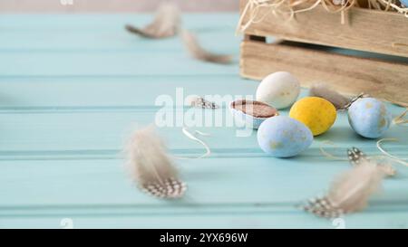 Easter eggs and feathers on a light blue wooden surface, with a wooden crate filled with straw in the background, creating a festive and rustic scene Stock Photo