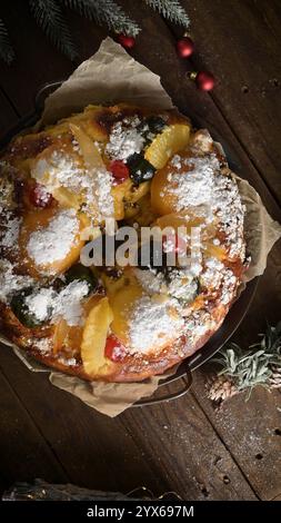 Festive fruitcake topped with powdered sugar, pineapple slices, cherries, and green candied fruits on a rustic wooden table with holiday decorations Stock Photo