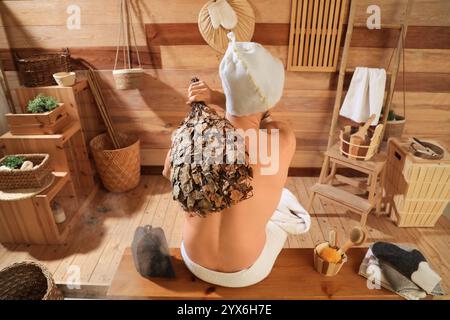 Young man with whisk relaxing on bench in sauna, back view Stock Photo