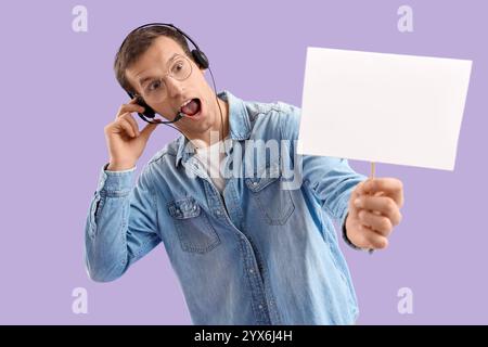 Shocked young man in headset with blank paper sign on lilac background Stock Photo