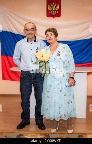 Russian senior couple celebrates their 50th golden wedding anniversary in a registry office. They stand together with medals and a bouquet of yellow r Stock Photo