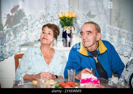 A 71-year-old Russian woman and her 76-year-old husband celebrate a festive occasion at a decorated table with food, flowers, and glasses. They look h Stock Photo