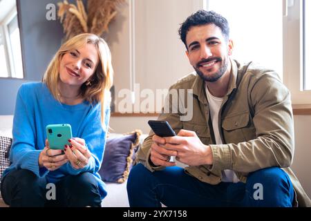 Couple sitting at home using smartphones, smiling and relaxed looking at camera Stock Photo