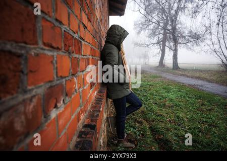 Sad lonely woman with hood standing by brick wall in fog. Loneliness and depression in own thoughts Stock Photo
