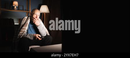 Elderly man watching TV at night holding remote control looking thoughtfully at screen with serious expression Stock Photo