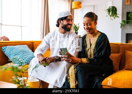 Happy beautiful young arab couple with emirati clothing together at home using cellphone for shopping online - Middle-eastern man and woman with tradi Stock Photo