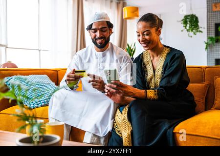 Happy beautiful young arab couple with emirati clothing together at home using cellphone for shopping online - Middle-eastern man and woman with tradi Stock Photo
