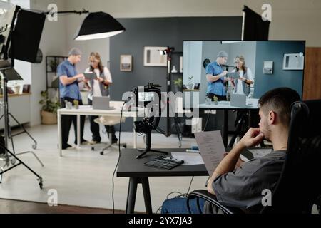 Unrecognizable man sitting at table and reading script while his coworkers discussing props and decorations for shooting space in background Stock Photo