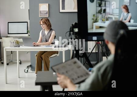 Elegant actress sitting at work desk and typing on keyboard while unrecognizable director with sketches in hand watching her Stock Photo