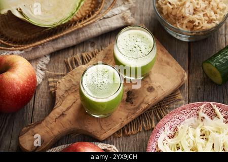 Fresh green cabbage and cucumber juice in two glass cups with red apples on a table Stock Photo