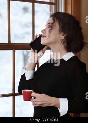 Woman with a cup of coffee talking on the phone by a window with a snowy winter scenery behind it. Apple iPhone. Stock Photo
