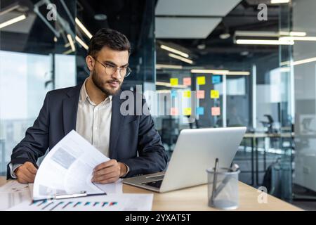 Mature man businessman sits at a desk, analyzing reports and working on a laptop. The professionally dressed individual is based in a bright, modern office with an organized workspace. Stock Photo